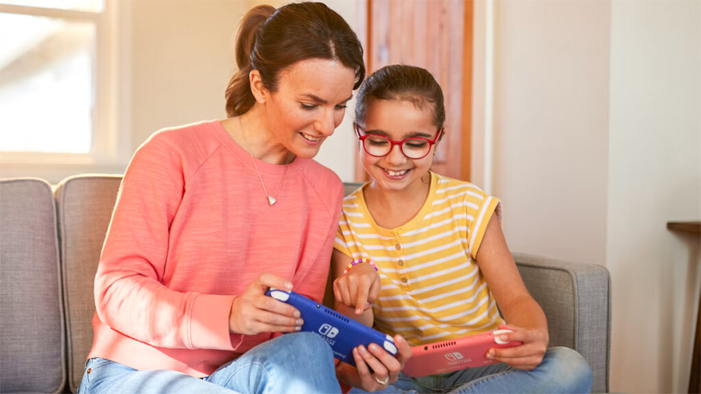 Mãe e filha jogam o Nintendo Switch Lite. Cada uma tem uma unidade do console em mãos, nas cores azul e coral