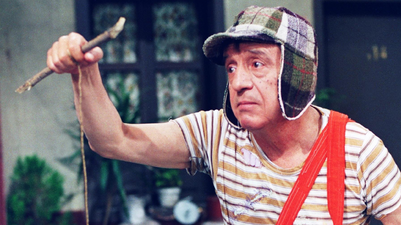 Um menino com um gorro xadrez predominantemente verde com abas caindo para proteger as orelhas, camisa listrada em branco e um amarelo bem claro desbotado