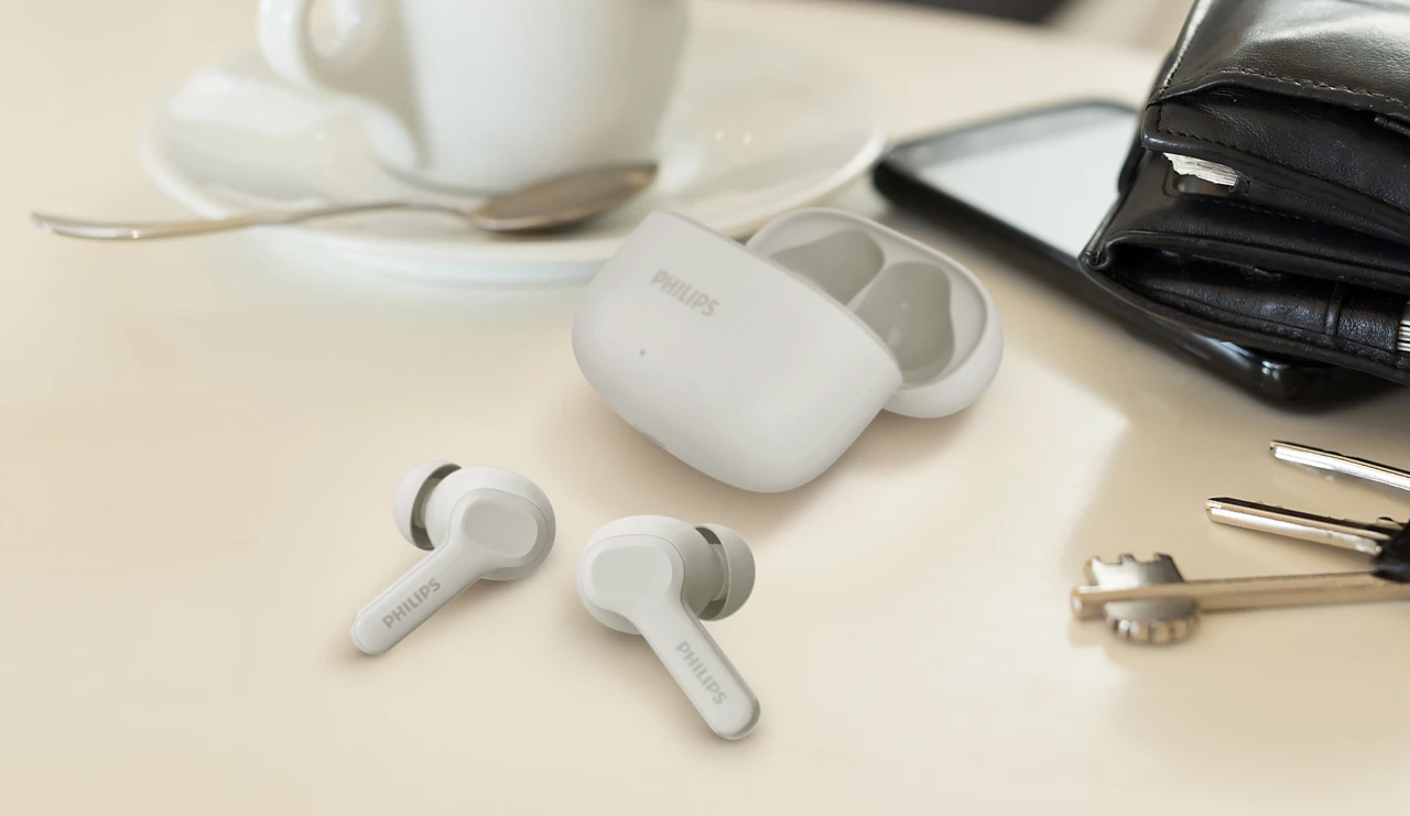 Fone de ouvido Philips TAT1109 na cor branca em uma mesa com xícara, chaves, carteira. Os fones estão fora do estjojo de carregamento, que está aberto acima deles