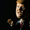 Foto de um boneco tipo bobble head de Donald Trump que parece fazer um gesto pedindo calma, com os braços meio abertos e as mãos espalmadas