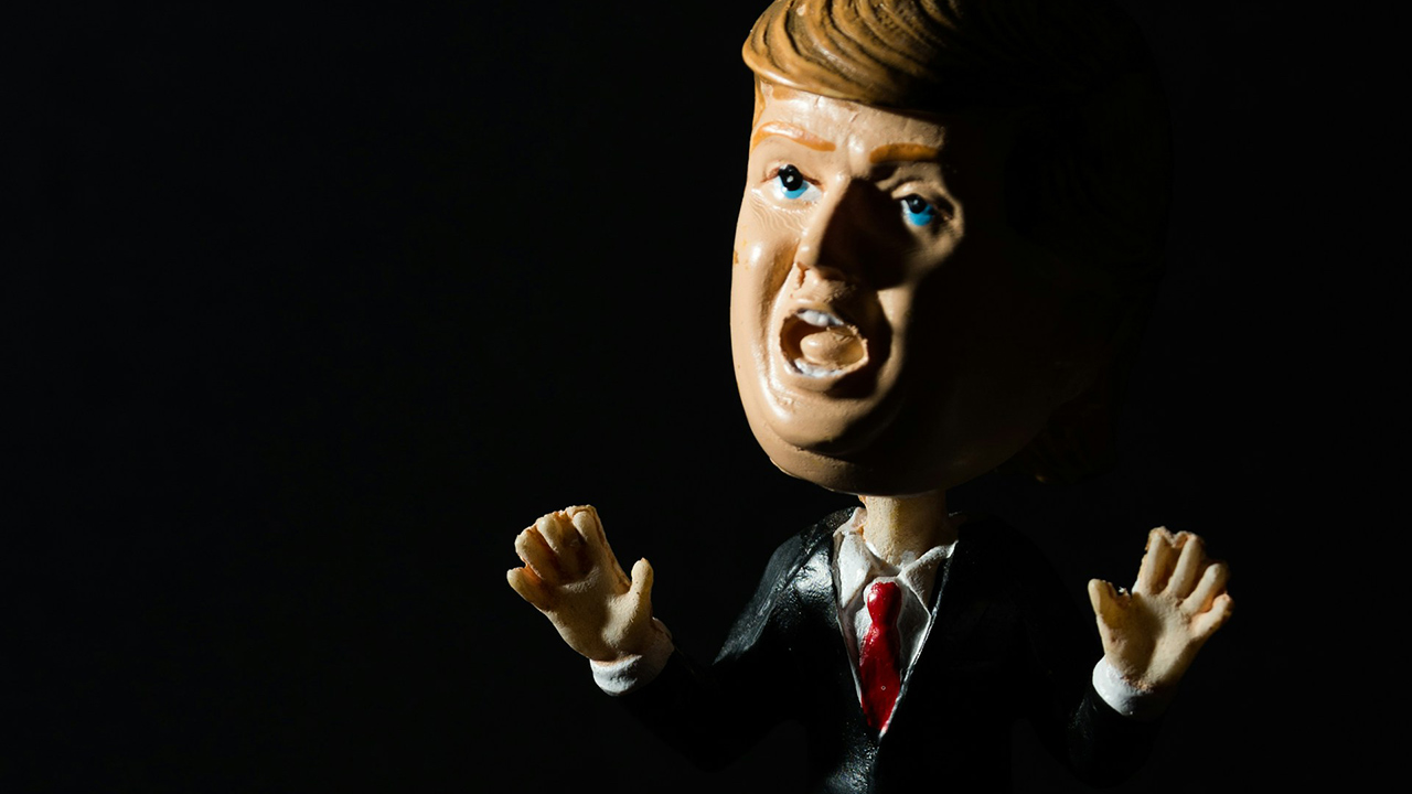Foto de um boneco tipo bobble head de Donald Trump que parece fazer um gesto pedindo calma, com os braços meio abertos e as mãos espalmadas
