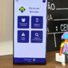 Foto de um celular na tela inicial do app Celular Seguro, com um playmobil de boné, celular pendurado e segurando um notebook e parte do globo terrestre