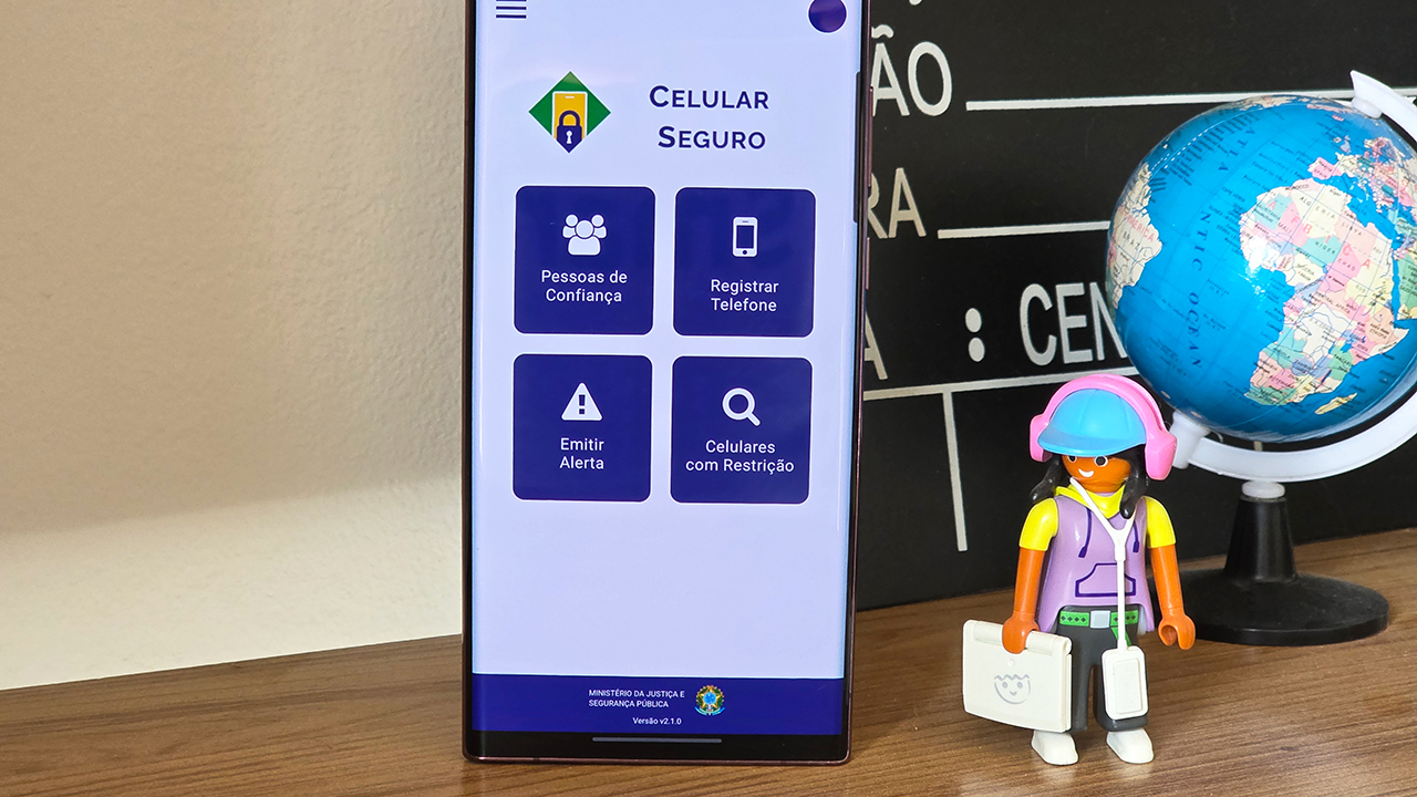 Foto de um celular na tela inicial do app Celular Seguro, com um playmobil de boné, celular pendurado e segurando um notebook e parte do globo terrestre