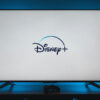 Uma televisão com a tela branca exibindo o logo do Disney+; a TV emite uma luz azulada na parede atrás dela