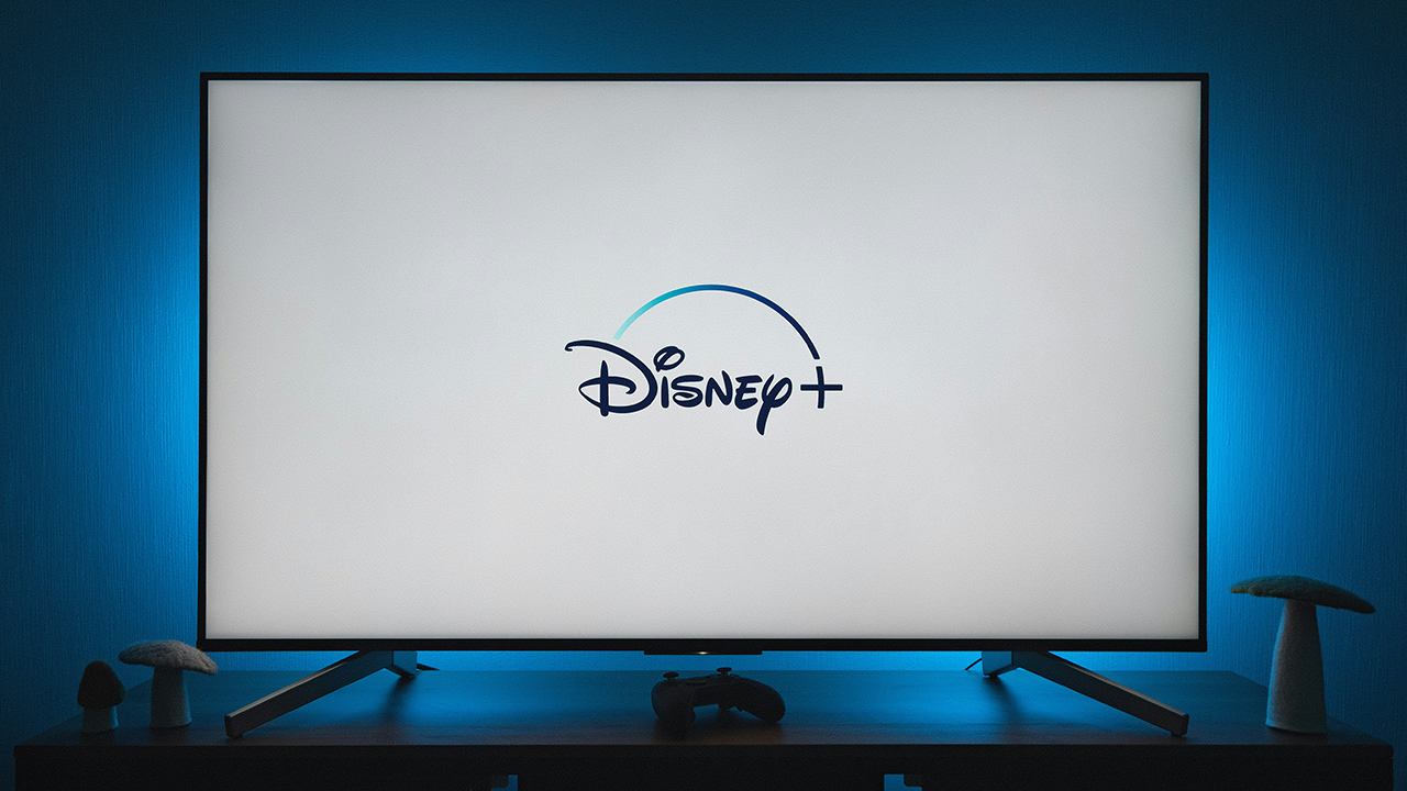 Uma televisão com a tela branca exibindo o logo do Disney+; a TV emite uma luz azulada na parede atrás dela