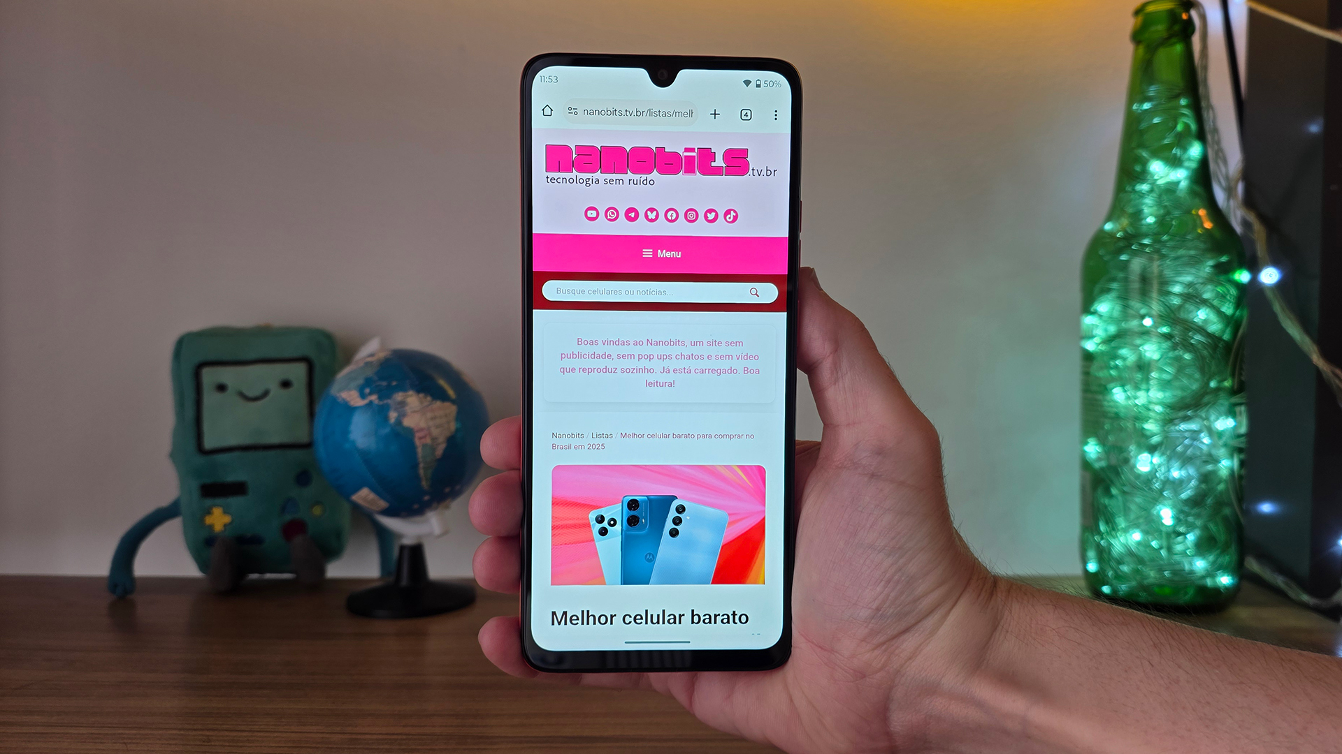 Uma pessoa segura um celular na página do Nanobits. Ao fundo, luzes e um globo desfocados