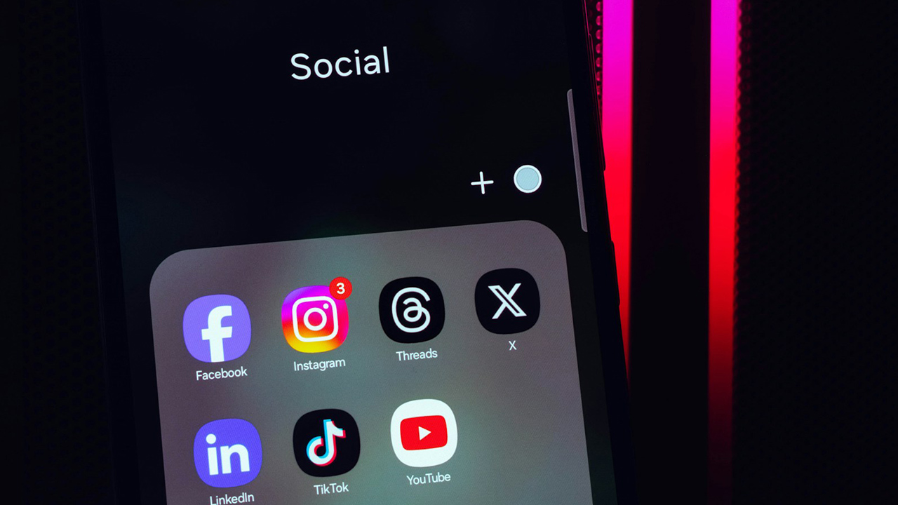 Foto de um celular na pasta 'Social' com os ícones de Facebook, Instagram, Threads, X/Twitter, LinkedIn, TikTok e YouTube
