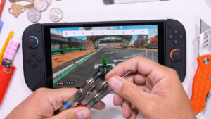 O Nintendo Switch 2 em uma bancada com estilete e outros objetos abrasivos e uma pessoa segura algumas pontas metálicas em frente à tela