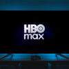 Uma TV com ologo da HBO Max na tela; a sala está escura e só tem uma luz traseira da televisão, na cor azul, deixando a silhueta de objetos como controle de videogame e o que parece uma luminária de cogumelo na mesa