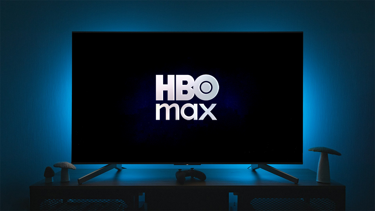 Uma TV com ologo da HBO Max na tela; a sala está escura e só tem uma luz traseira da televisão, na cor azul, deixando a silhueta de objetos como controle de videogame e o que parece uma luminária de cogumelo na mesa