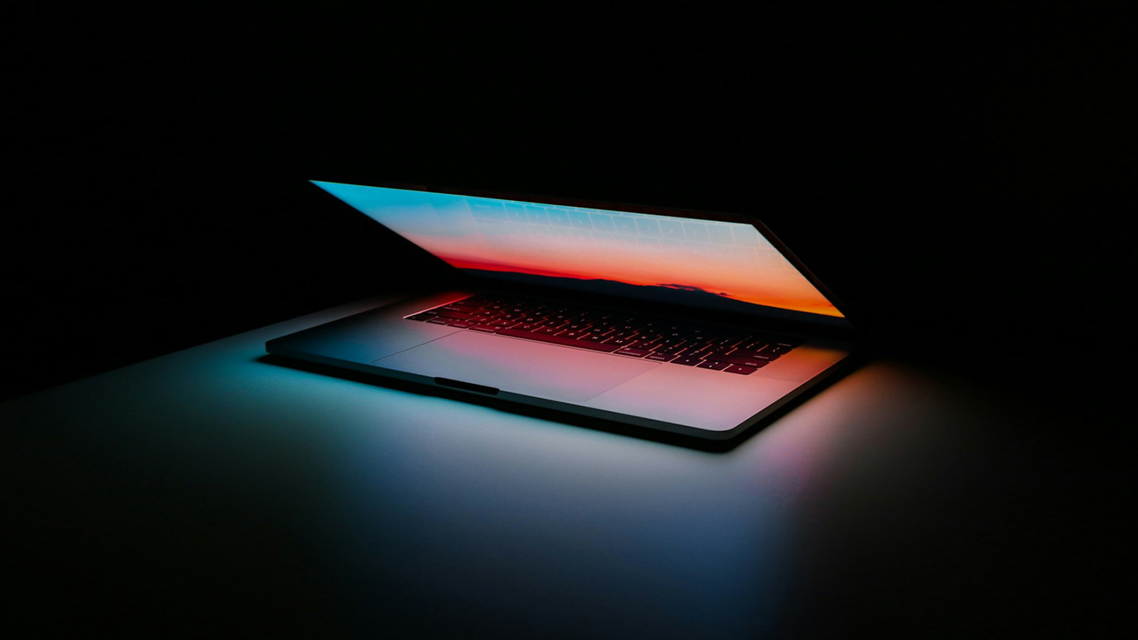 Um MacBook ligeiramente aberto em um local escuro, apenas a tela do notebook ilumina seu teclado