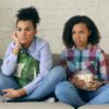 Duas mulheres assistem a um filme sentadas no sofá com um balde de pipoca